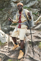 Portrait of content young Afro American hiker with exercise mat in backpack holding walking poles against rocky mountain