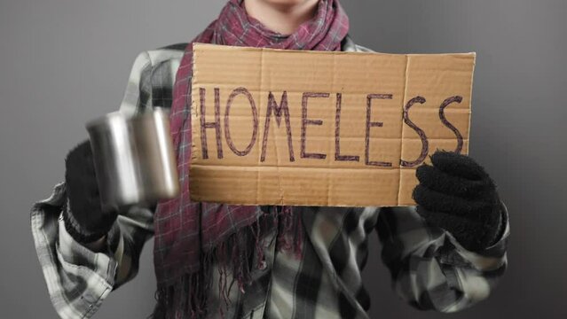A untidy beggar woman in dirty clothes and gloves begging and holds a cardboard sign with the inscription homeless. Dark background. Close up. The concept of helping vagrants.