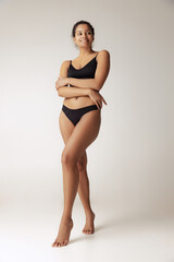Full-length portrait of young slim woman posing in black underwear isolated over grey background