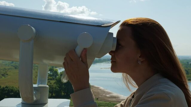 Young woman looking through big street binoculars at beautiful scenery. Tourist girl using viewing machine for observing nature panorama with river, forest, meadow. Lady watching through telescope