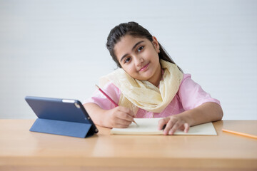 portrait cute Indian girl writing on notebook and online learning class from tablet, education...