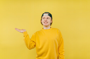 Positive young man in a yellow sweatshirt stands on a yellow background with a smile on his face and looks away with a smile on his face and shows his hand on copy spacee