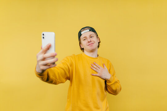 Joyful guy in a cap and casual clothes takes a selfie on the smartphone camera on a yellow background and laughs.