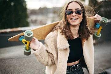 Gordijnen Stylish young woman holding skateboard on shoulders on roof © WellStock