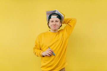 Positive young male student in casual clothes and cap standing with notebook in hand and pen, looking at camera and smiling ironically