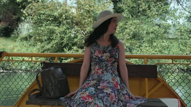 A beautiful young lady in a straw hat and dress is sitting and admiring nature. The concept of summer vacations and river excursions.