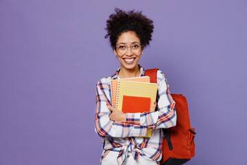 Young smiling satisfied girl woman of African American ethnicity student in shirt backpack glasses hold books isolated on plain purple background. Education in high school university college concept.