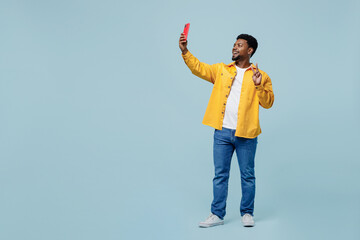 Full body fun young man of African American ethnicity wear yellow shirt doing selfie shot on mobile cell phone post photo on social network show v-sign isolated on plain pastel light blue background. - Powered by Adobe