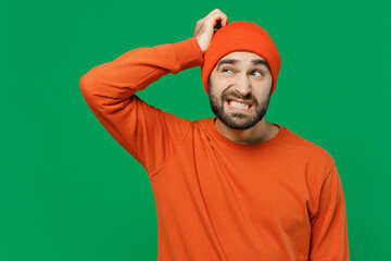 Fototapeta premium Young sad puzzled mistaken confused caucasian man 20s wearing orange sweatshirt hat look aside hold scratch head say oops isolated on plain green background studio portrait. People lifestyle concept.