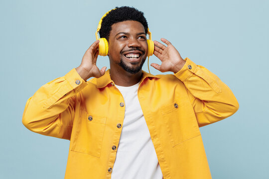 Young fun cool man of African American ethnicity 20s wear yellow shirt headphones listen to music look aside isolated on plain pastel light blue background studio portrait. People lifestyle concept