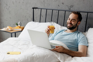 Young freelancer man in blue t-shirt lying in bed hold use work on laptop pc computer drink juice...