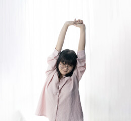 Standed Little Asian young Glasses Girl stretches oneself in front of light white curtain background.
