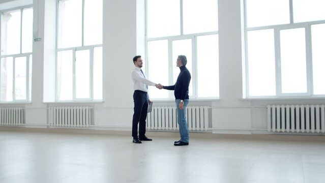 Businessman shakes hands with realtor in an empty rental space, contract for mortgage, side view. Realtor meets client in room purchased with mortgage