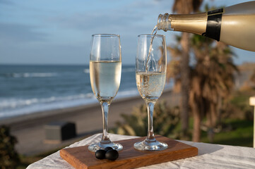 Pouring of Spanish cava sparkling wine is glasses with view on blue sea and sandy beach, Costa del Sol vacation destination, Andalusia, Spain