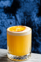 A Glass of a yellow cocktail with white foam big ice cube decorated with orange chips on the blue blurred background