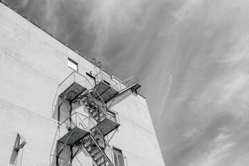 Black and White Image of a fire escape on the side of tall building and wispy clouds in the background