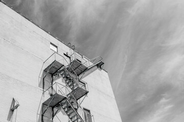 Black and White Image of a fire escape on the side of tall building and wispy clouds in the...