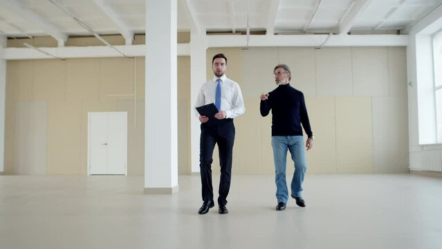 Businessman and manager walk around an empty warehouse discussing business, front view. Businessman walks through an empty room with realtor discussing lease agreement