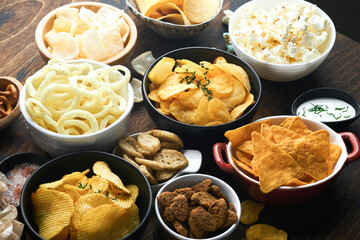 Unhealthy food or snacks. All classic potato snacks with peanuts, popcorn and onion rings and salted pretzels in bowl plates on old wooden background. Unhealthy food for figure, heart, skin, teeth.