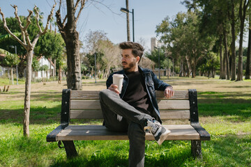 Fototapeta premium Enjoying in the park, young handsome man sitting on the bench and crossed his legs, he is holding a coffee mug in his hand, Young adult enjoying a spring day