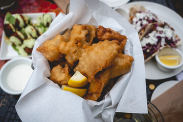 Closeup of basket with lemon and fried tempura vegetables, fish, chicken and meat in a table with more food ('falafel', 'tacos') in the day