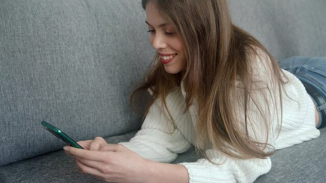 A beautiful young girl lies on the couch with a modern smartphone in her hands and looks through the content on the screen and smiles.