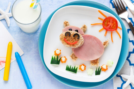 Fun food for kids - cute smiling hippo sandwich shaped of toast bread and ham decorated with carrots, cucumbers and tomatoes for lunch and served with milk