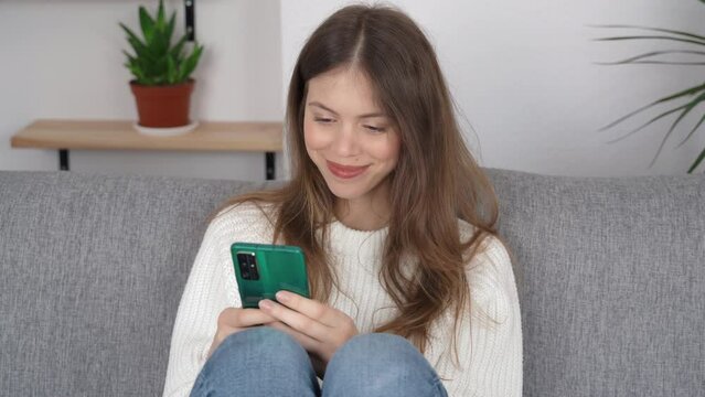 A young girl is sitting in a relaxed position on the sofa with her legs up and a phone in her hands, she is typing something on the screen and smiling. Closeup view.