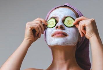Beautiful young woman in facial cosmetics mask putting cucumber slices on eyes.