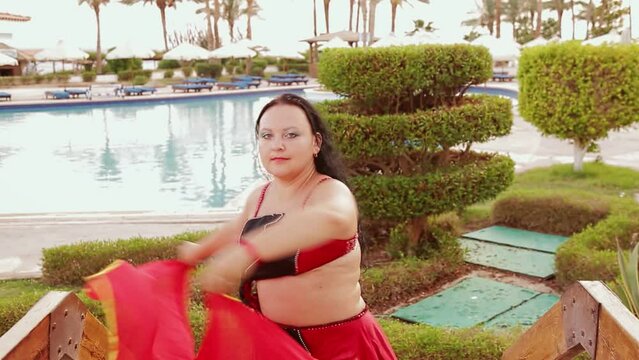 A brunette woman in a red oriental dance costume with a shawl dances by the pool. Medium plan