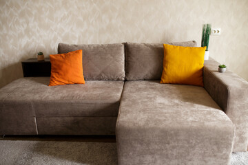 a new light gray sofa next to a beige wall. two orange pillows on the sofa and houseplants near the sofa.