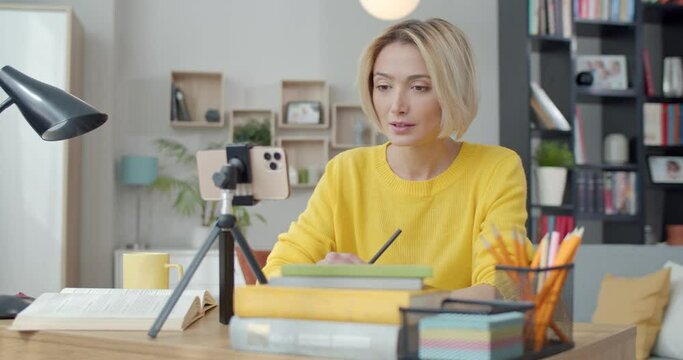 Caucasian young woman having video call on modern smartphone while studying at home. Pleasant female blonde sitting at desk, talking and taking notes.