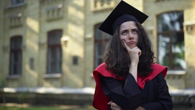 Thoughtful young beautiful woman in mortarboard cap and graduation toga standing at university campus on the right looking away. Portrait of slim charming Caucasian student thinking about future