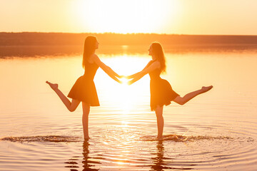 Two sisters in black dresses dance on the water, holding hands in the contrasting rays of the sunset.
