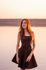 Close-up portrait of young woman in black dress against the sunset sky. Woman walks on the seashore