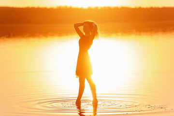 Bright, sunny Silhouette of young woman in airy black dress in the water of lake with sky reflection