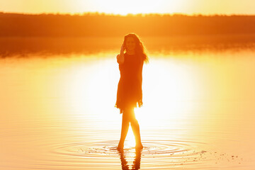 Fototapeta premium Bright, sunny Silhouette of young woman in airy black dress in the water of lake with sky reflection