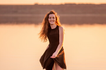 Close-up portrait of curly, happy dancer in airy black dress at sunset. Young woman in the water