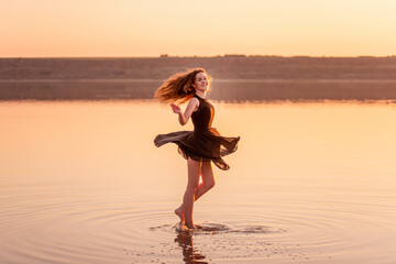 Bright, sunny Silhouette of young curly dancer in airy black dress in the water of lake with sky