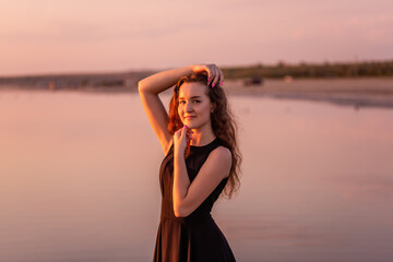 Fototapeta na wymiar Close-up portrait of curly, happy dancer in airy black dress at sunset. Young woman in the water