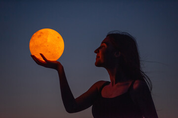 A young woman holds the full moon in her hands against the backdrop of a red sunset. Astrology