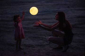 Young mother plays with little daughter. A woman throws a full moon lamp to a girl from hand to hand
