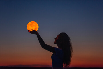 A young woman holds the full moon in her hands against the backdrop of a red sunset. Astrology