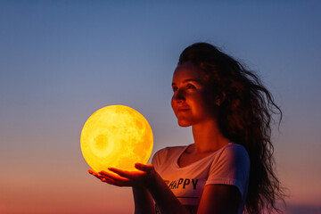 A young woman holds the full moon in her hands against the backdrop of a red sunset. Astrology