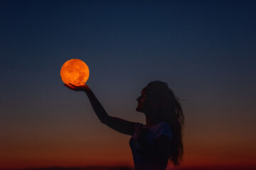 A young woman holds the full moon in her hands against the backdrop of a red sunset. Astrology
