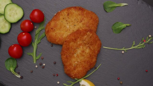 High angle view of beautifully cooked golden brown vegan schnitzel surrounded by a mosaic of garnishes tomatoes, cucumber, arugula, peppercorns, lemon on a gray slate platter