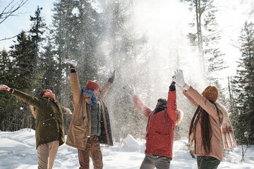 Group of young playful men and women enjoying spending time outdoors on winter day throwing snow in...