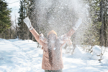 Cheerful young Black woman with long hair spending sunny winter day in local forest throwing snow in the air
