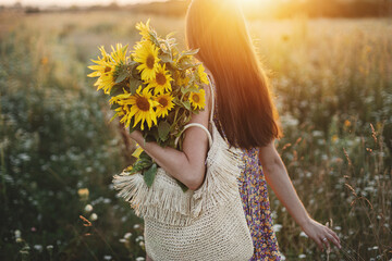 Beautiful woman holding sunflowers in warm sunset light in meadow. Tranquil atmospheric moment in countryside. Stylish young female in floral dress walking with sunflowers in summer evening field