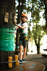 A cheerful boy in an orange helmet has fun in the rope park at sunset in summer